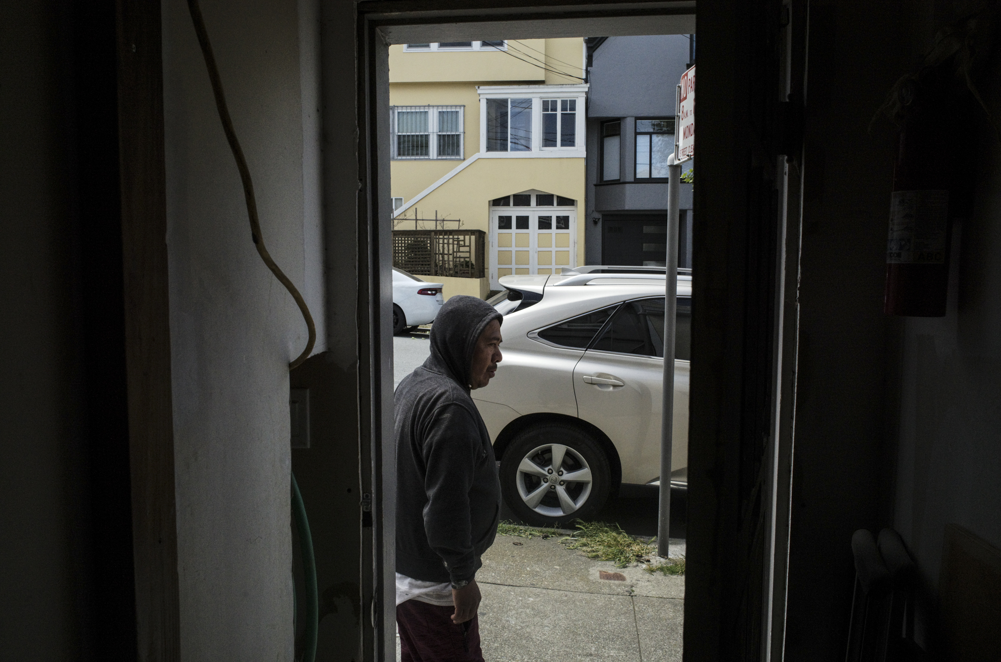 A middle-aged Latino man outside of his house, photographed from inside the house, with a car parked on the street outside his house.