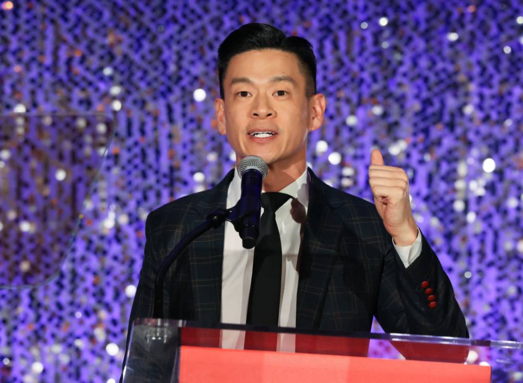 A young Asian person wearing a black suit and tie speaks into a mic at a clear podium on a stage.