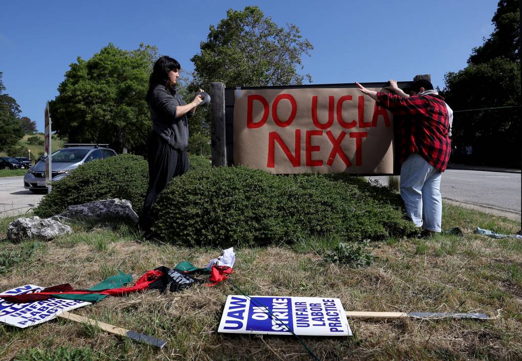 Two people affix a large brown sign with red text reading 'Do UCLA next' to a school sign outdoors near a busy street.