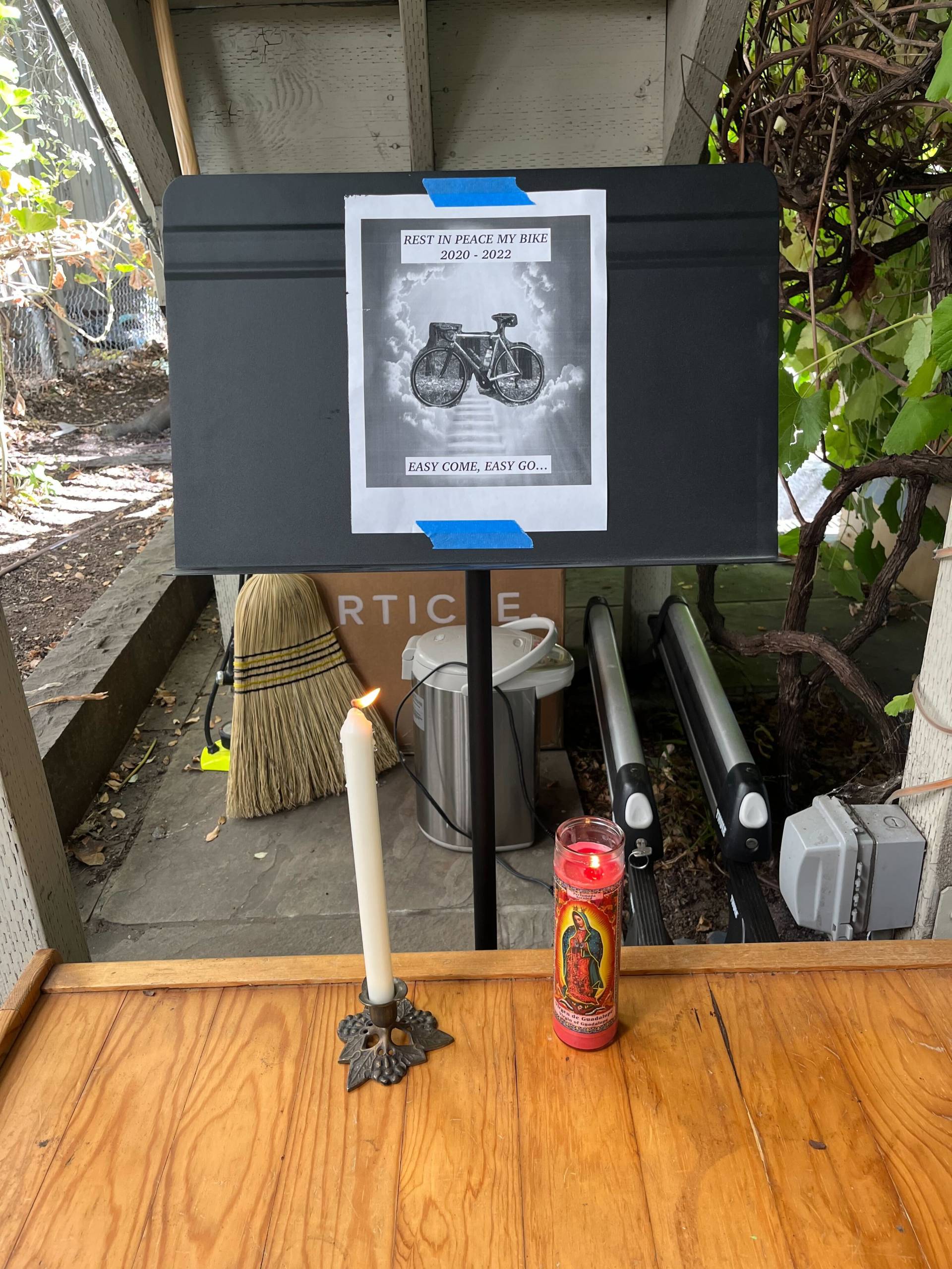 In a backyard, there is a music stand. On the music stand, there is a piece of paper with a photo of a bicycle printed on it. In front of the music stand, there are two candles.