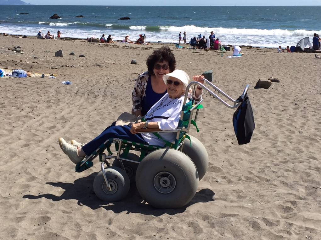 A person wearing a white shirt and white cap sits on a beach wheelchair with the ocean visible in the background. Another person stands next to her in the photo.