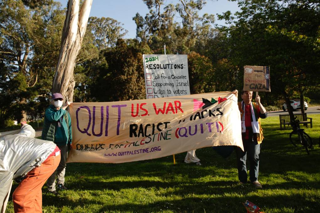 a small group holds up signs and a banner that says 'quit US war' in a park
