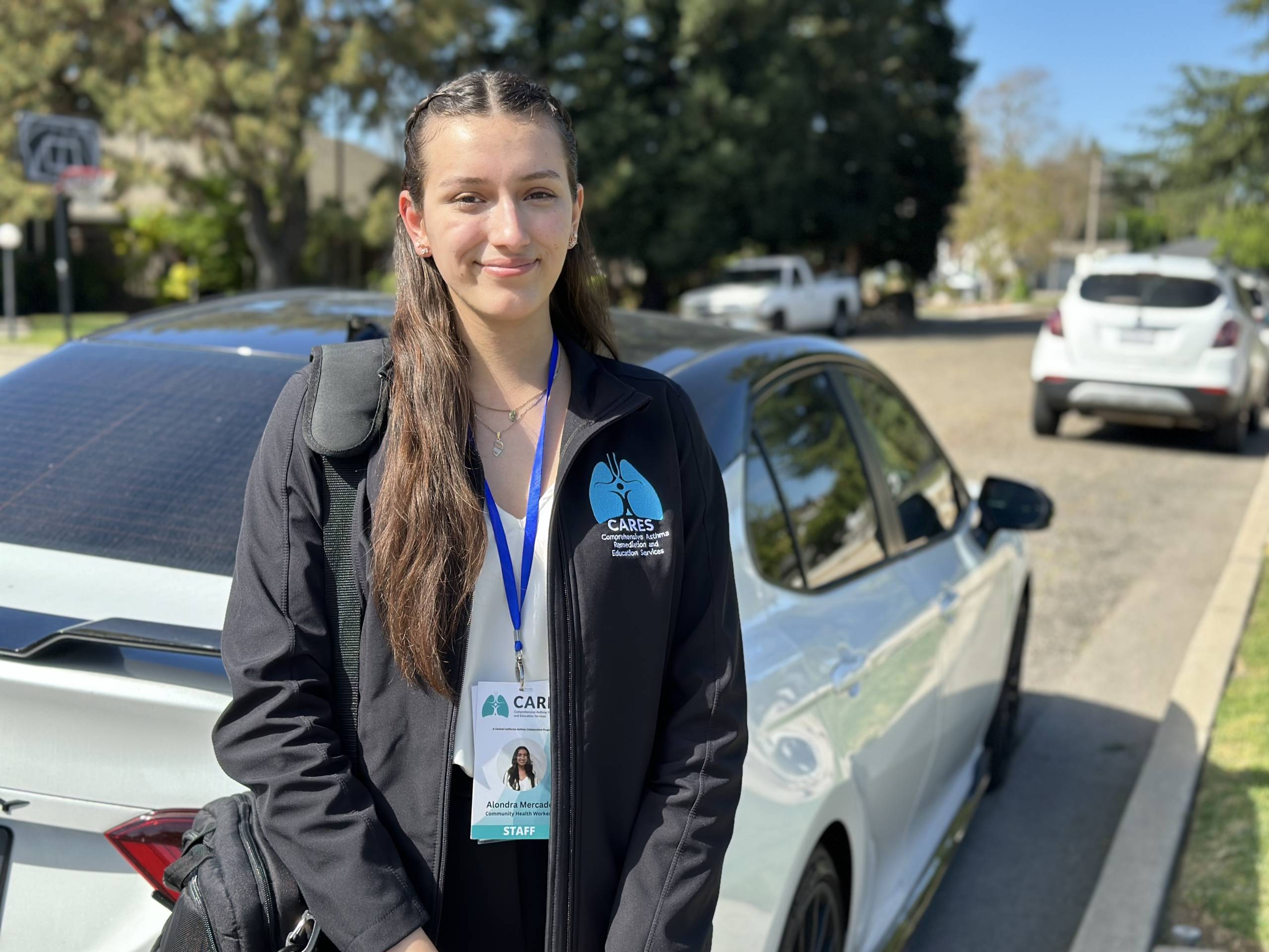 A woman wearing a black jacket, white shirt and lanyard stands behind a vehicle parked on the street.