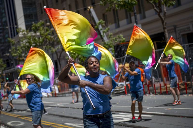 A group of flag twirlers holding rainbow flags perform on a street.