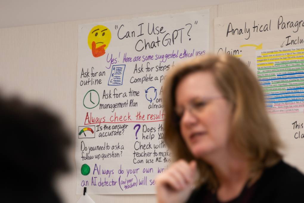 A sign that reads "Can I Use ChatGPT?" is posted on the wall behind a white woman with glasses.