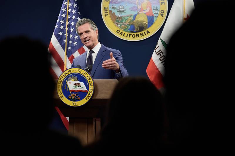 A white man wearing a business suit stands behind a podium with flags on both sides and a state logo above him while gesturing with his left hand.