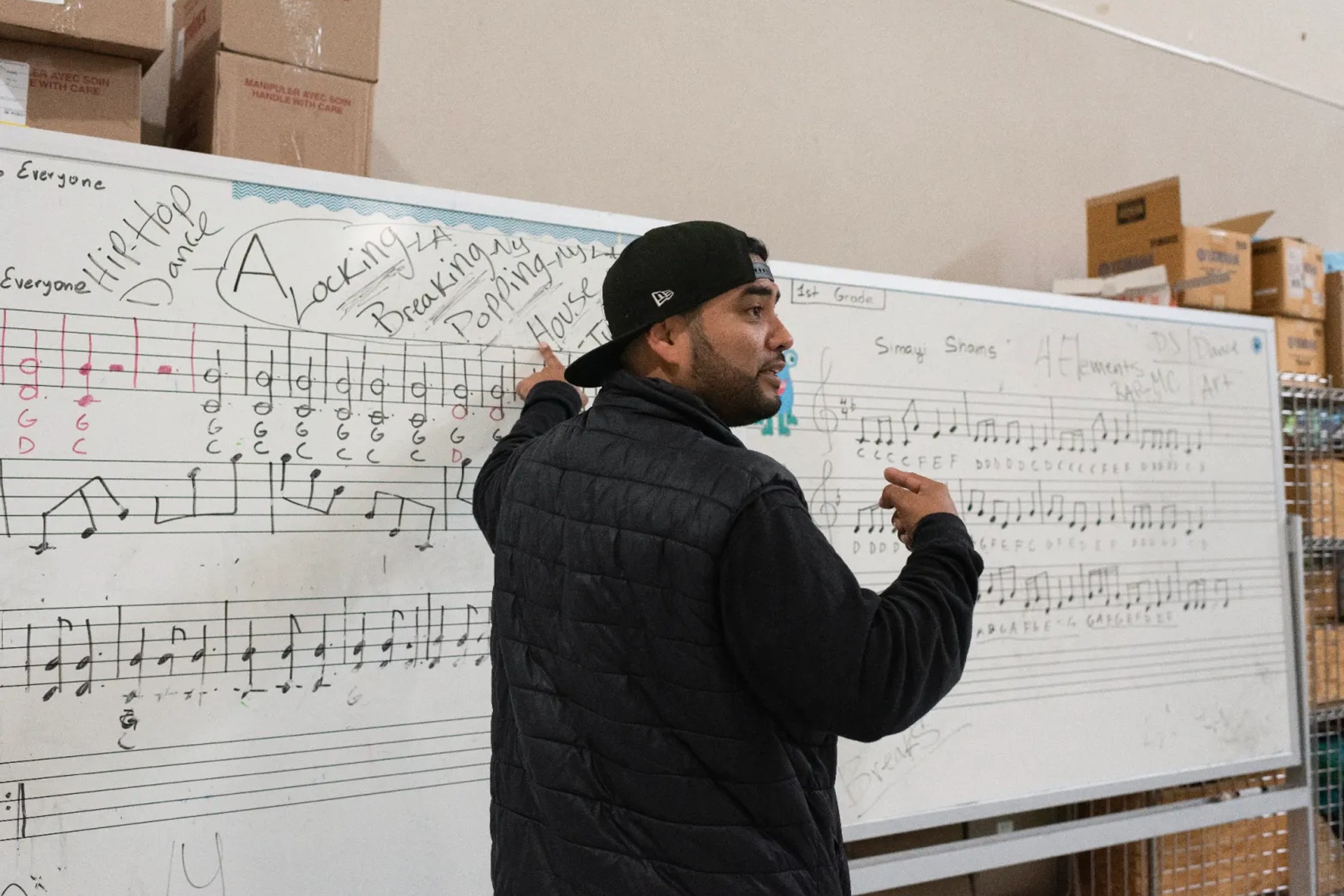 A man with a backward cap on stands in front of a whiteboard. Musical scales are written on the board. 
