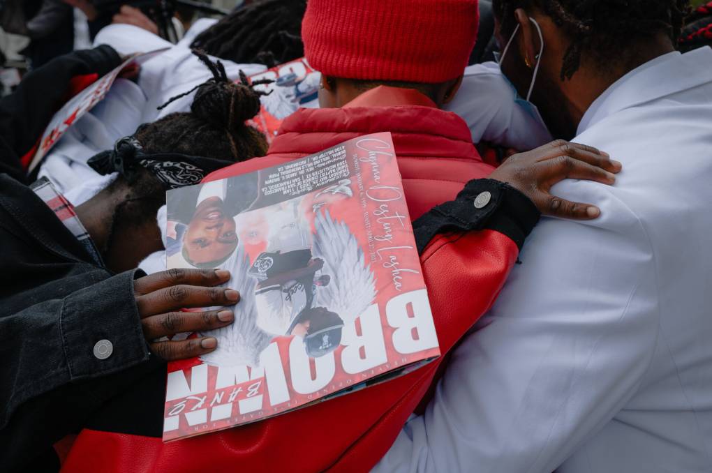 A group of people embrace each other at a funeral. One holds a pamphlet that says, "Brown" in big white letters.