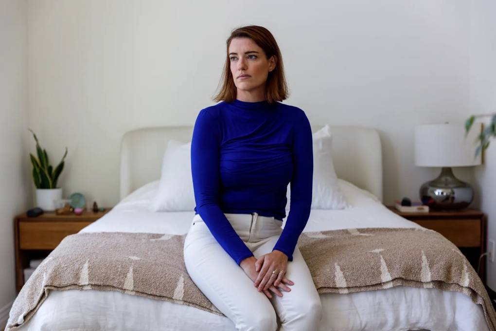 A young woman in a blue shirt and white pants sits on the foot of a bed.