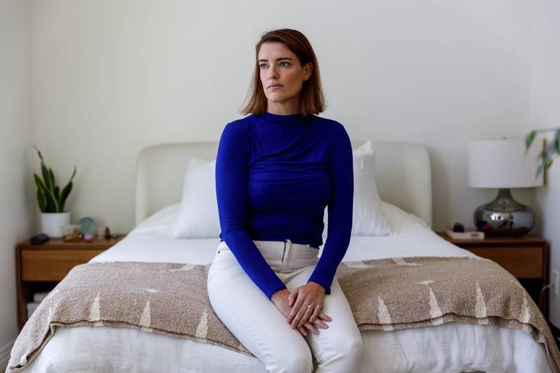 A young woman in a blue shirt and white pants sits on the foot of a bed.