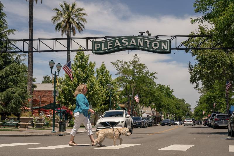 A white woman wearing a blue shirt and white pants walks a dog across the street with a sign that says "Pleasanton" above her.