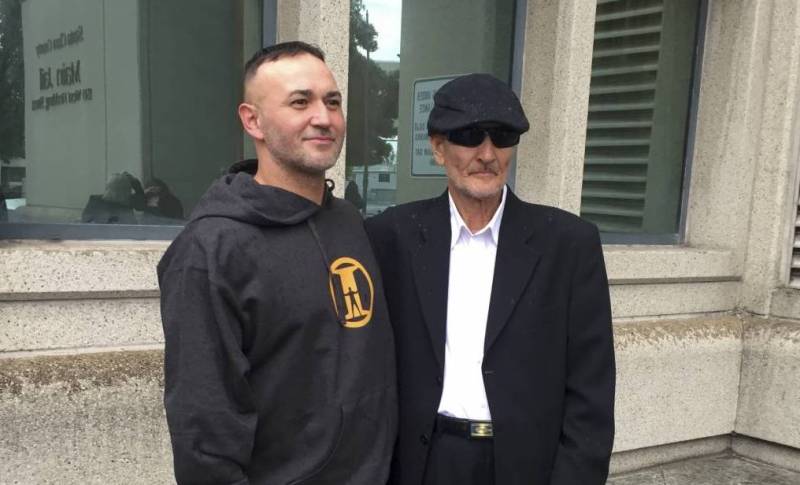 A younger man poses outside next to an older man (his father).