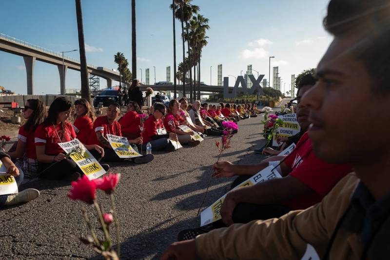 Protesters sit on the ground, holding signs, outside of Los Angeles International Airport — with LAX in large letters behind them.