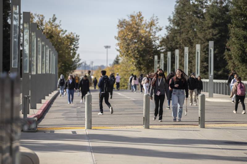 Various groups of people walk on a bridge leading to a college campus.