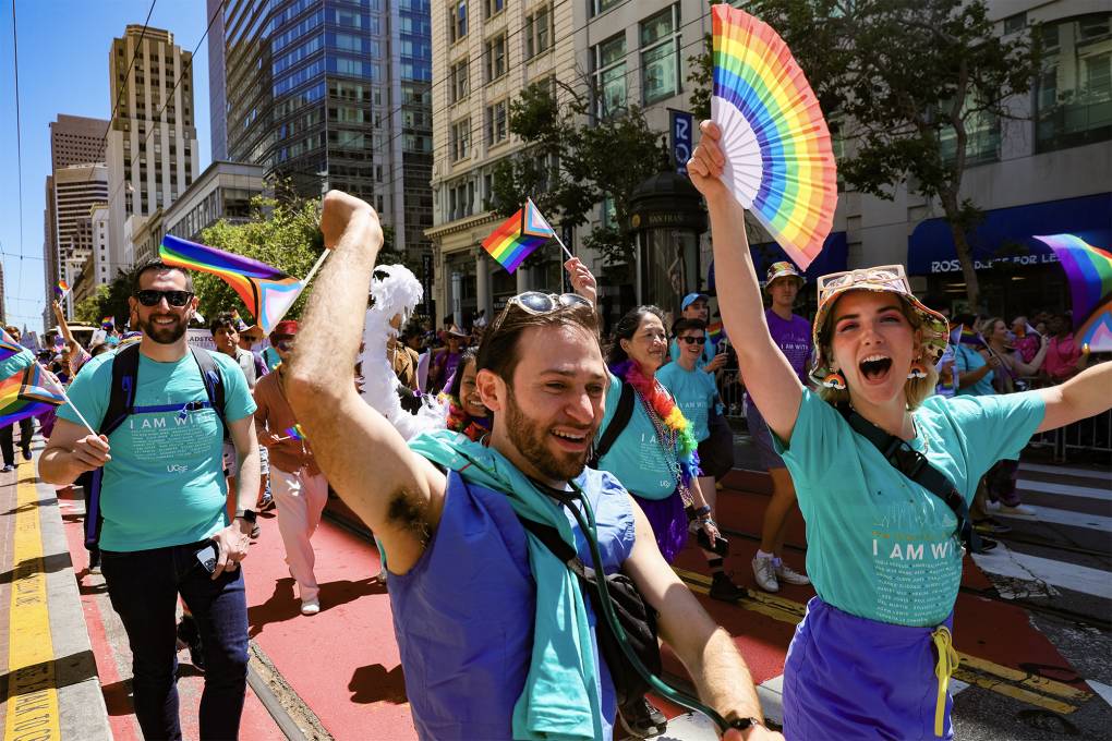 A group of people wearing light turquoise shirts wave rainbow flags and fans while walking down the street.
