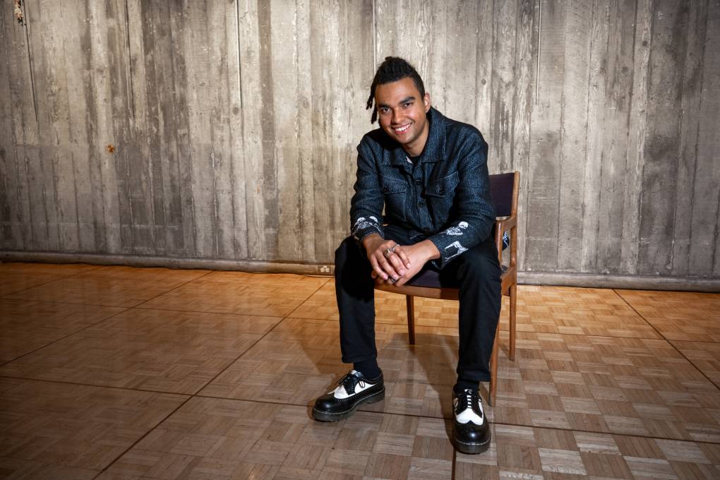 A young Black man sits on a chair with his hands together.