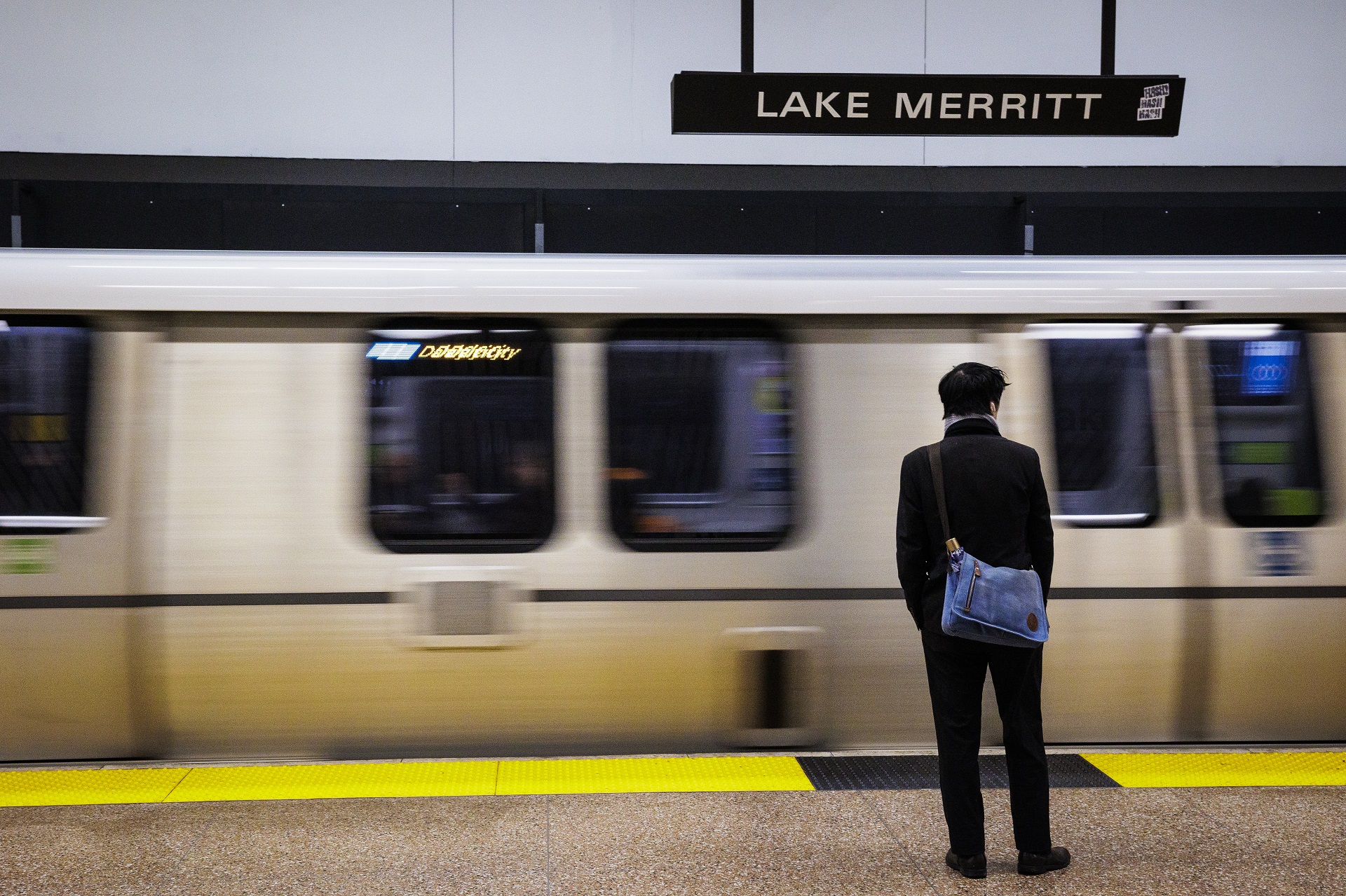 A man stands at a train platform as a train leaves the station. A sign above says "Lake Merritt."