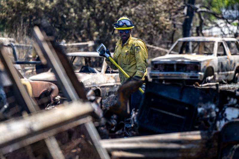 A man wearing fire safety equipment and holding a tool walks past charred remains of vehicles.