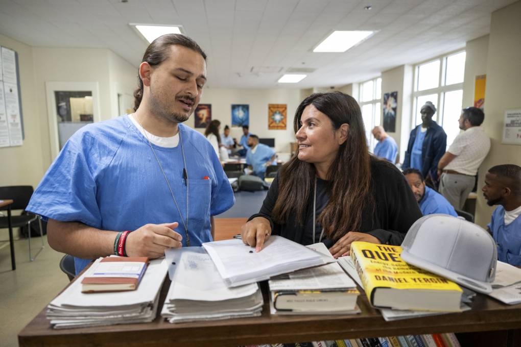 A man with long, braided hair in a blue prison jumpsuit stands next to a much shorter woman in front of a table of books.