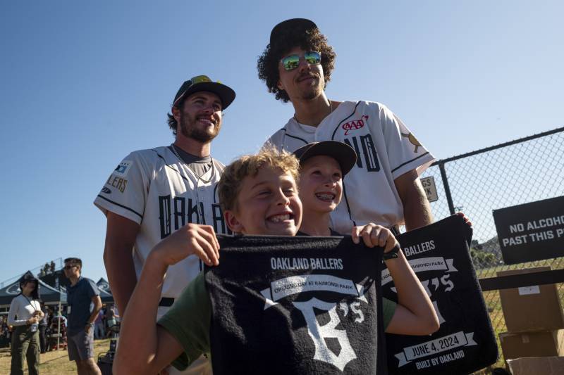 Two men dressed in baseball uniforms stand in front of two young boys holding up fabric that reads "Oakland Ballers."