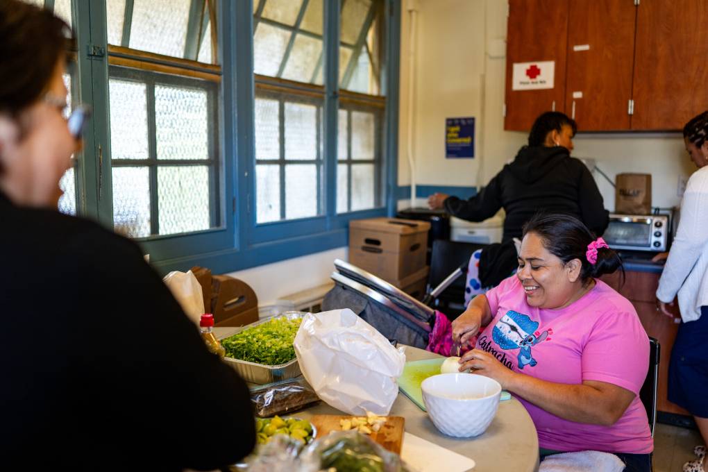 A woman wearing glasses and a black shirt talks to a woman wearing a pink t-shirt in a kitchen, cutting food with two other people in the background.