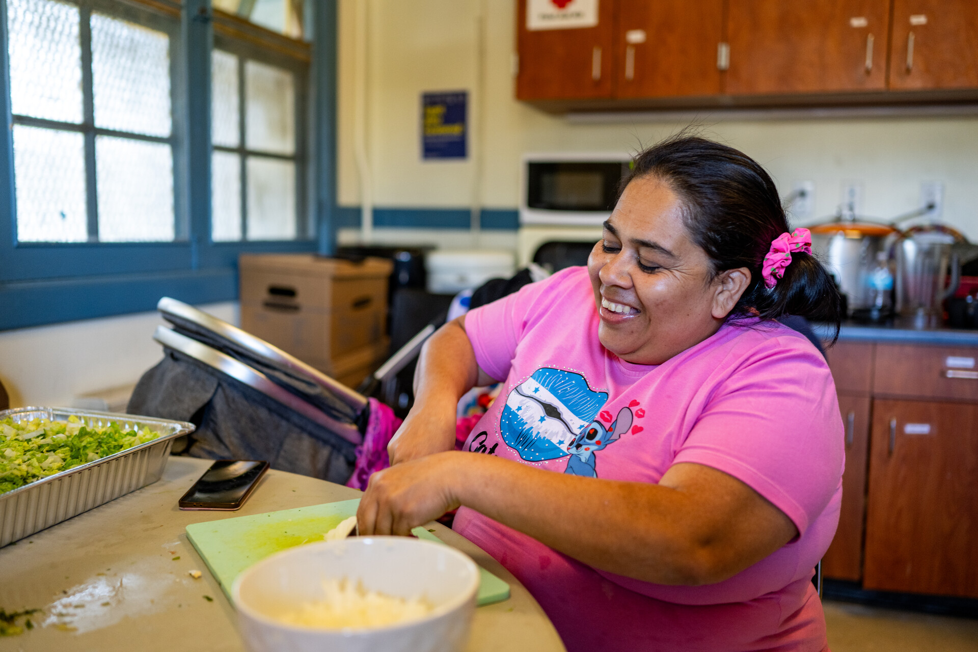 A woman wearing a pink t-shirt cuts food on a cutting board in a kitchen.