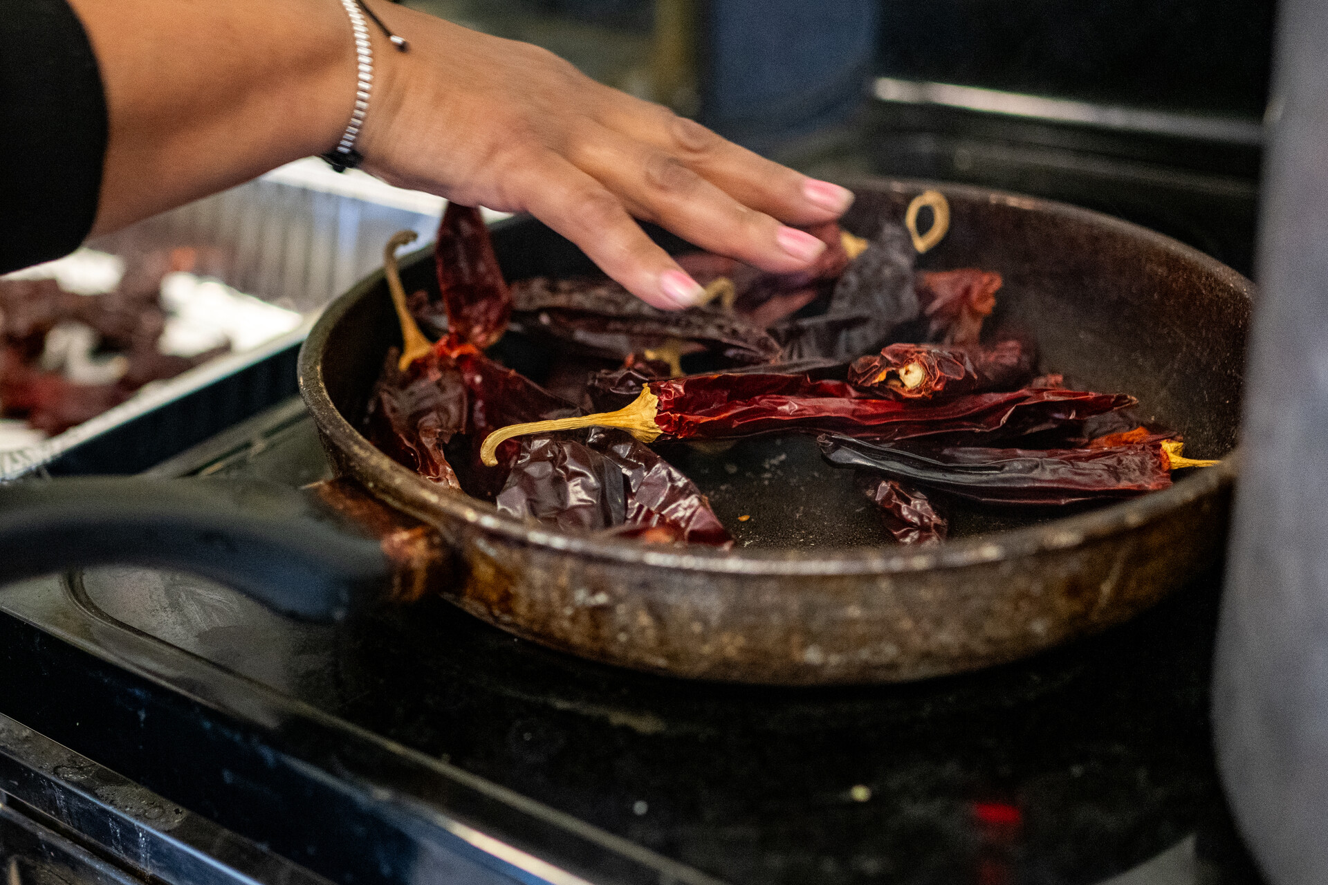 A hand touches chiles in a pan on a stovetop.