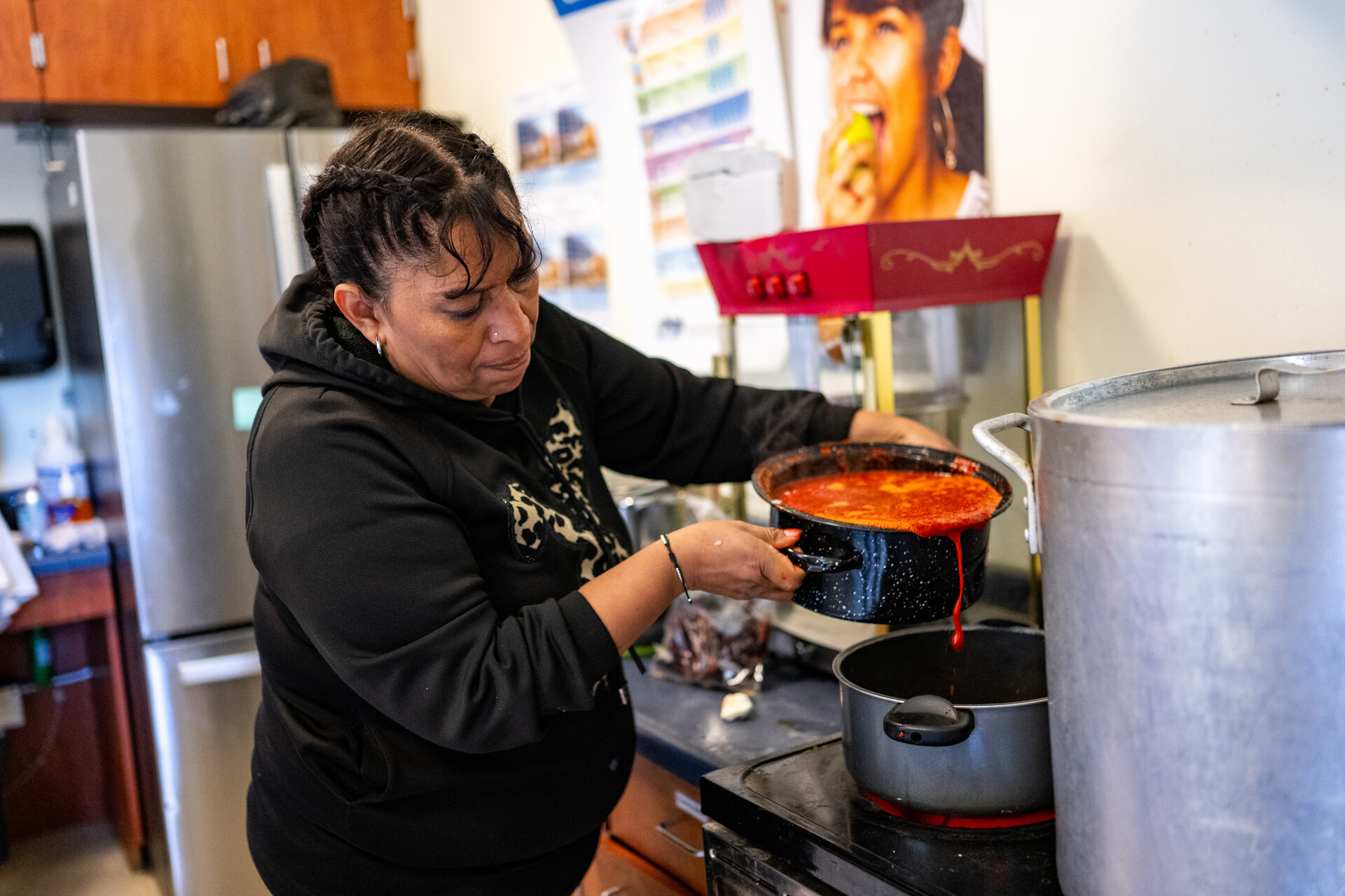 A woman wearing a black hooded sweatshirt pours soup into a large pot in a kitchen.