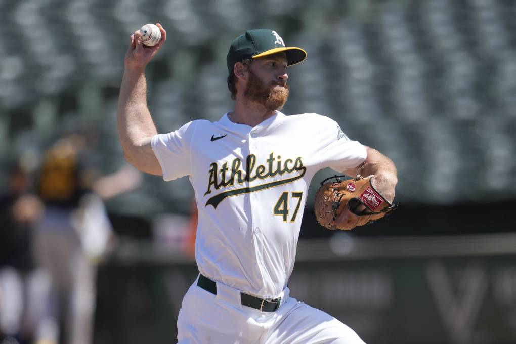 A man in a baseball uniform with "Athletics" across his chest throws a baseball.