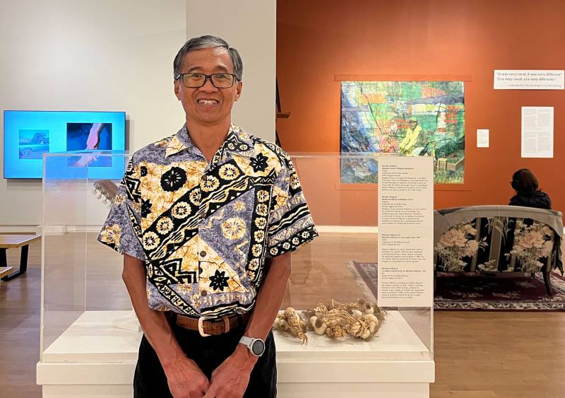 A man wearing glasses and a shirt with exotic designs stands in front of a display of old garlic.
