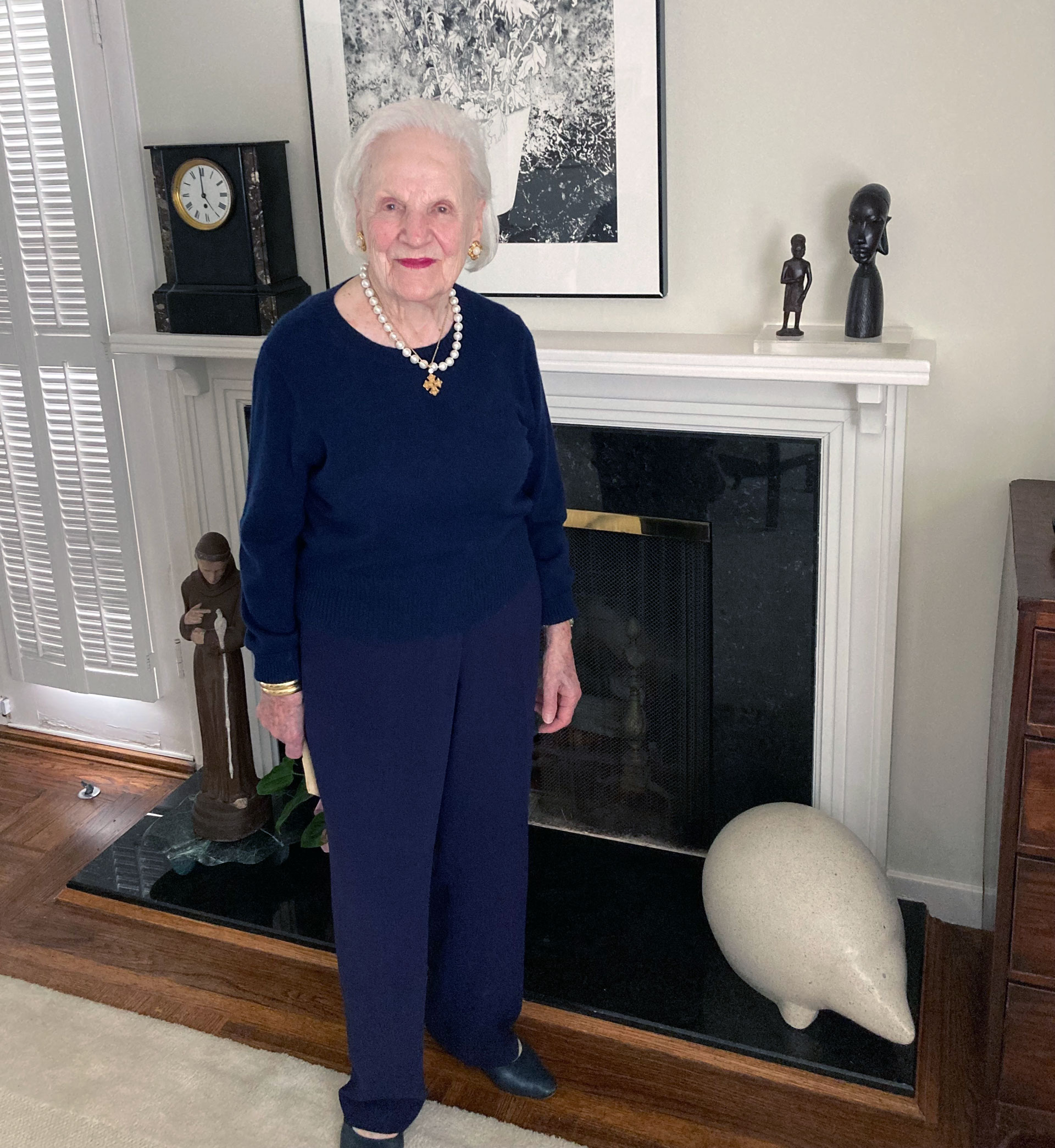 Older woman in a navy blue pants suit poses by a fireplace.