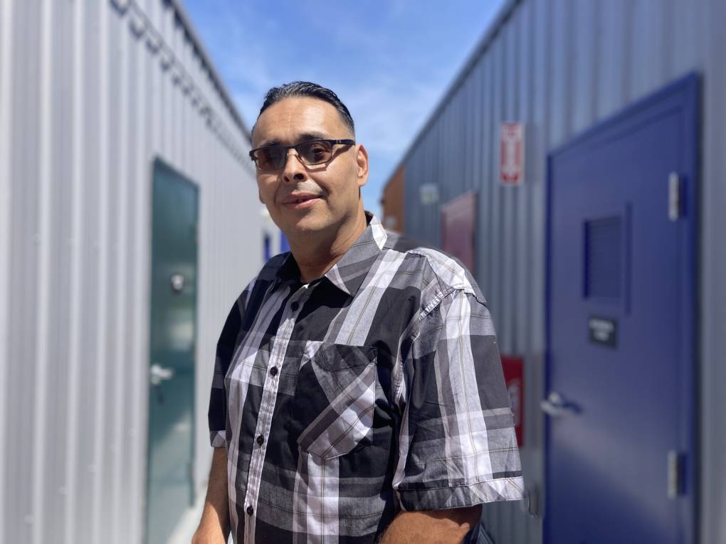 A young man with sunglasses and a plaid shirt poses for a photo standing in a walkway between two rows of grey tiny houses.