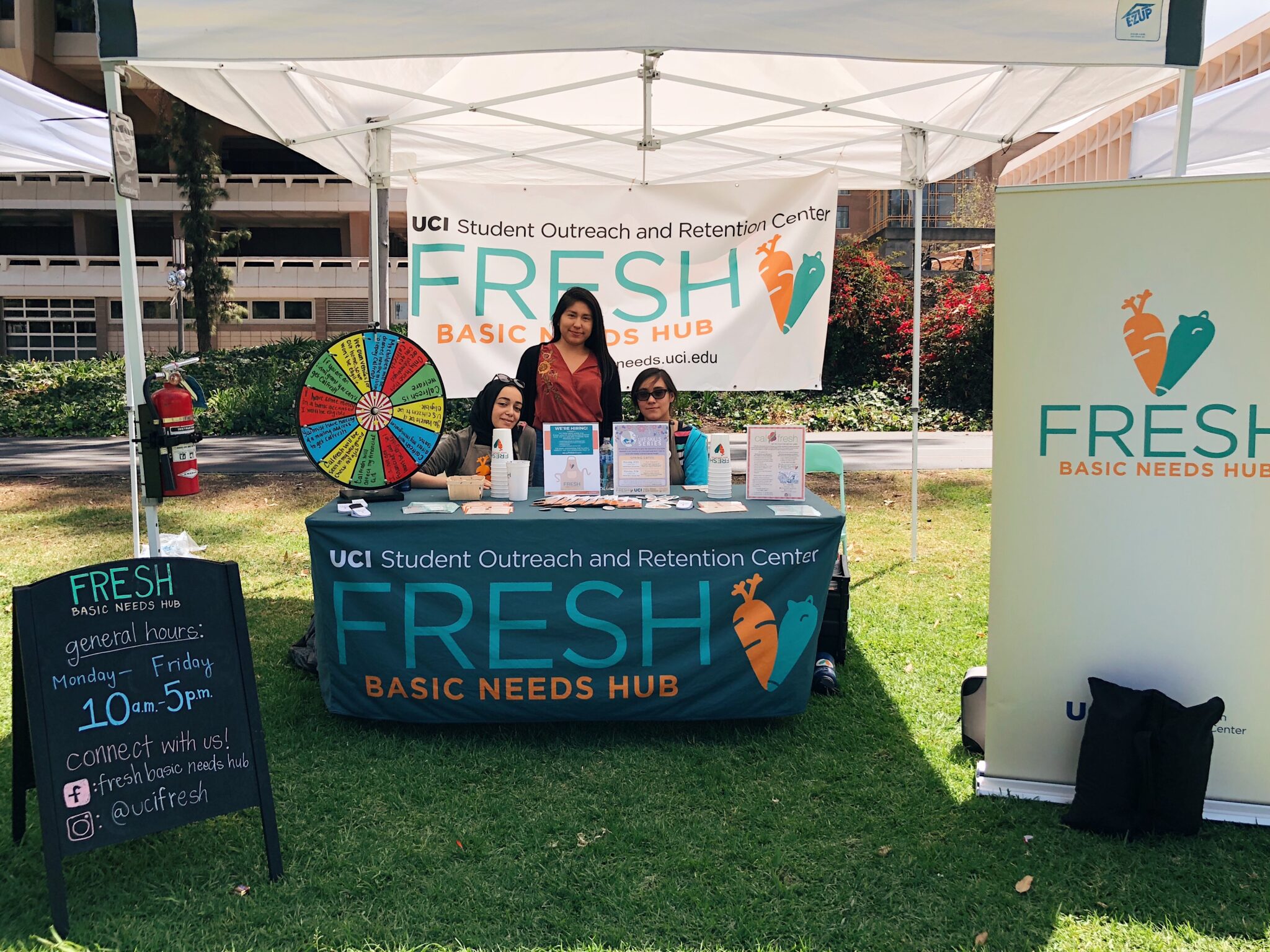 Three young women sit and stand behind a table filled with brochures and papers next to a colorful wheel with a banner and sign that reads "FRESH Basic Needs Hub."