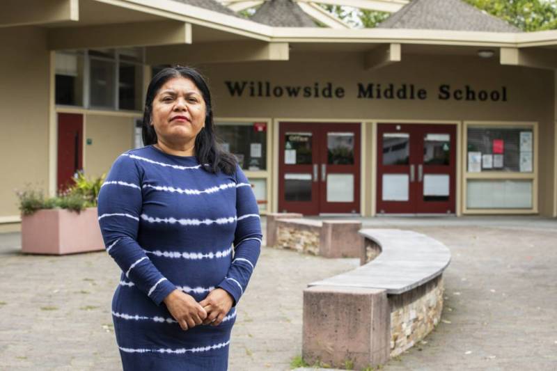 Una mujer latina lleva puesta un vestido azul y ve directamente a la cámara. Ella está parada enfrente de la escuela Willowside Middle School.