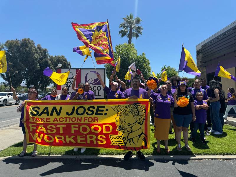 Fast food workers from KFC and Taco Bell march behind a huge red and yellow banner that reads, "San Joe fast food workers." Some demonstrators hold flags and signs.