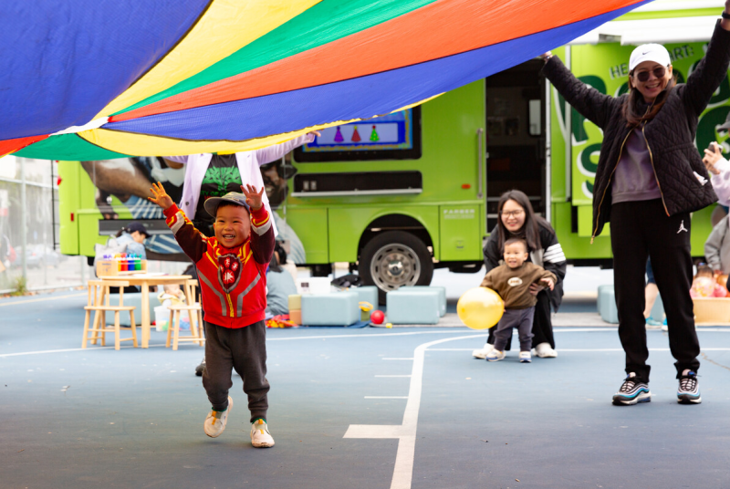 A small Asian boy runs, smiling, under a multicolored parachute, as his mother and another younger boy watch.