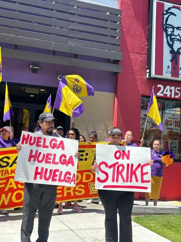 Two workers at a Taco Bell-KFC stand outside the location protesting unsafe working conditions. They hold signs that read, "Huelga, Huelga, Huela" and "On Strike."