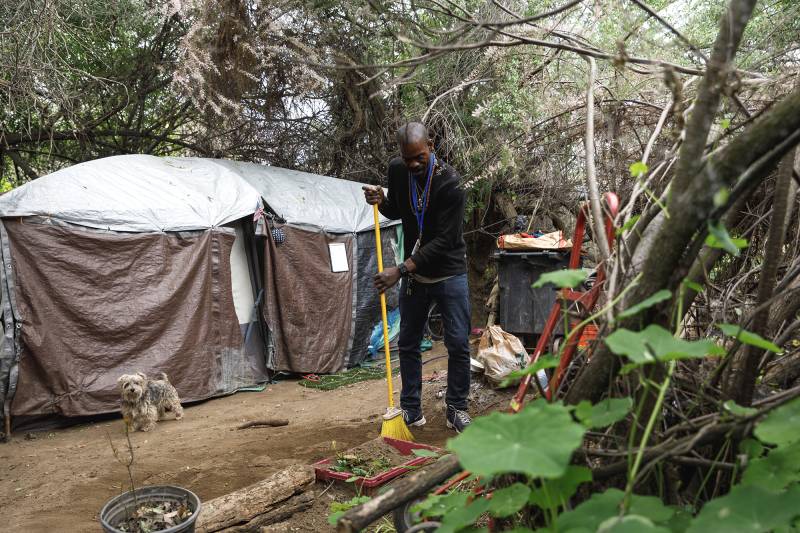 A Black man wearing dark clothing holds a broom in front of an encampment surrounded by trees.