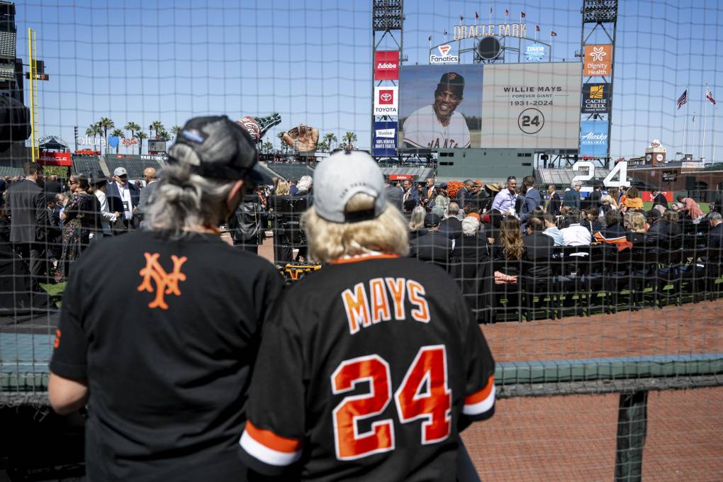 A man and a woman wearing black and orange baseball jerseys look at a crowd at a baseball stadium.