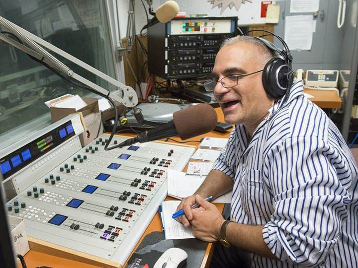 A man wearing a striped button down shirt with glasses and headphones speaks into a microphone with an audio control panel in front of him.