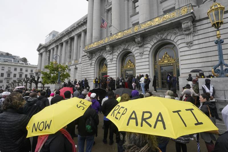 Several people stand outside of a building looking at people standing in front. Two people hold yellow umbrellas that say "SF Reparations Now."