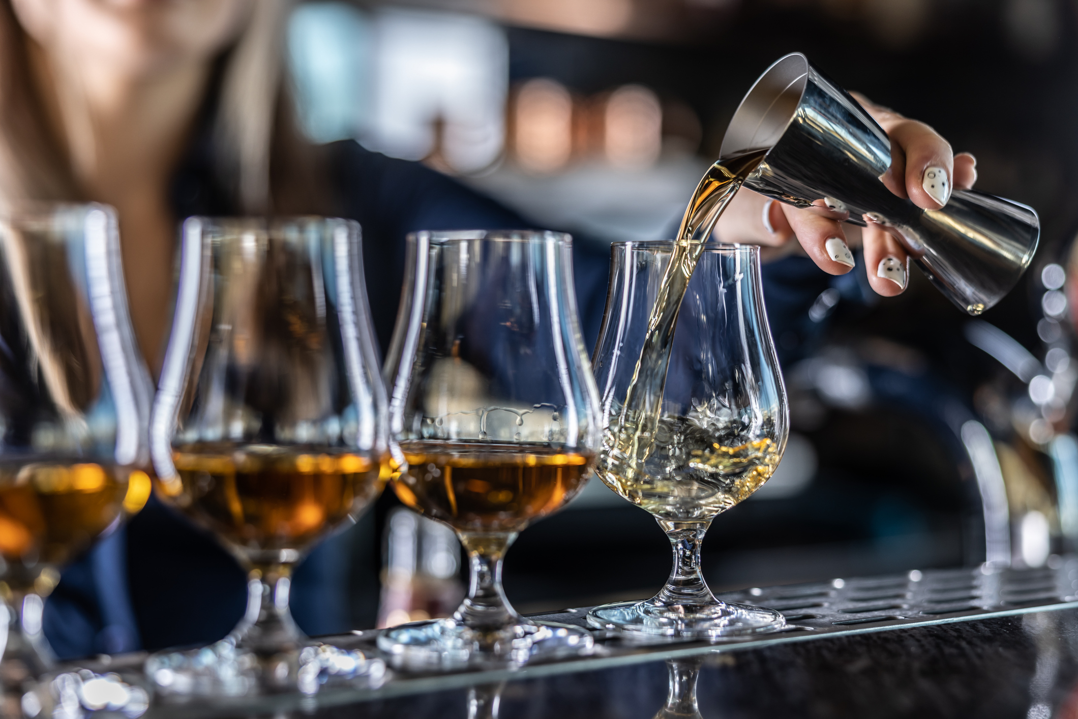 A young woman pours a brown liquor from a steel jigger into the glass at a bar.