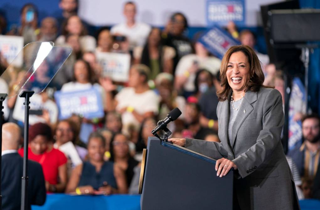 A woman smiles from a podium with several people in the background.