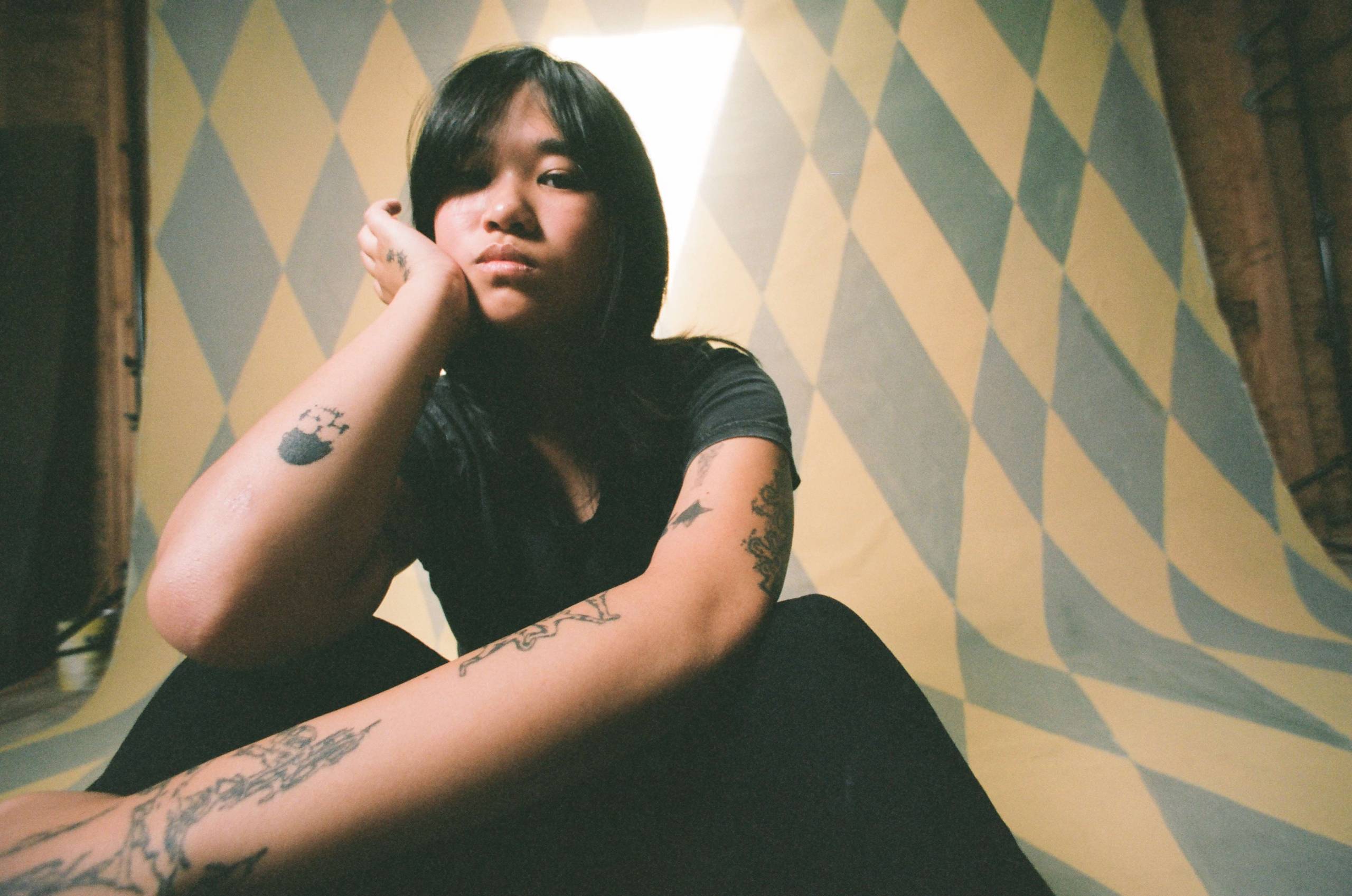 An Asian woman with tattoos wearing a dark short sleeve shirt rests her head on her right hand.
