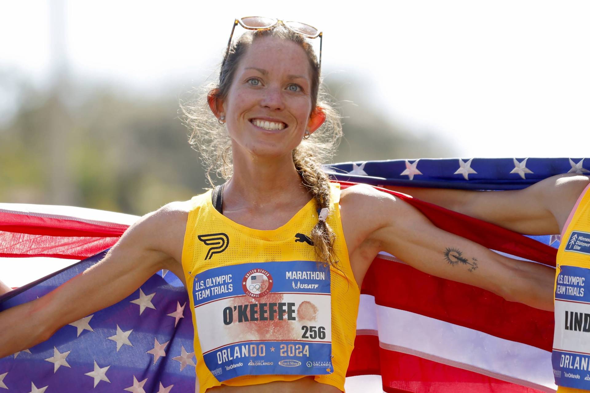 A female marathon runner smiles broadly with her arms outstretched, partially wrapped in an American flag. She has a patch of blood on the front of her shirt.