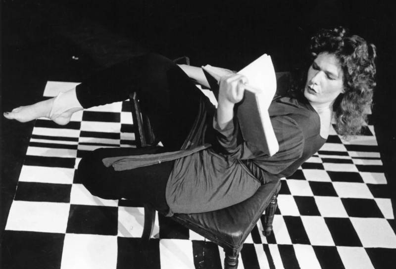 Black and white image of a woman reading a book. She is sitting crooked on a chair with her knees pulled up and feet in the air. Beneath her is a striking checkerboard floor.