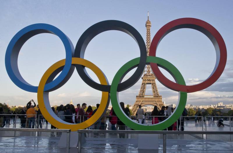 Olympic rings statue in front of the Eiffel Tower with people crowded around.