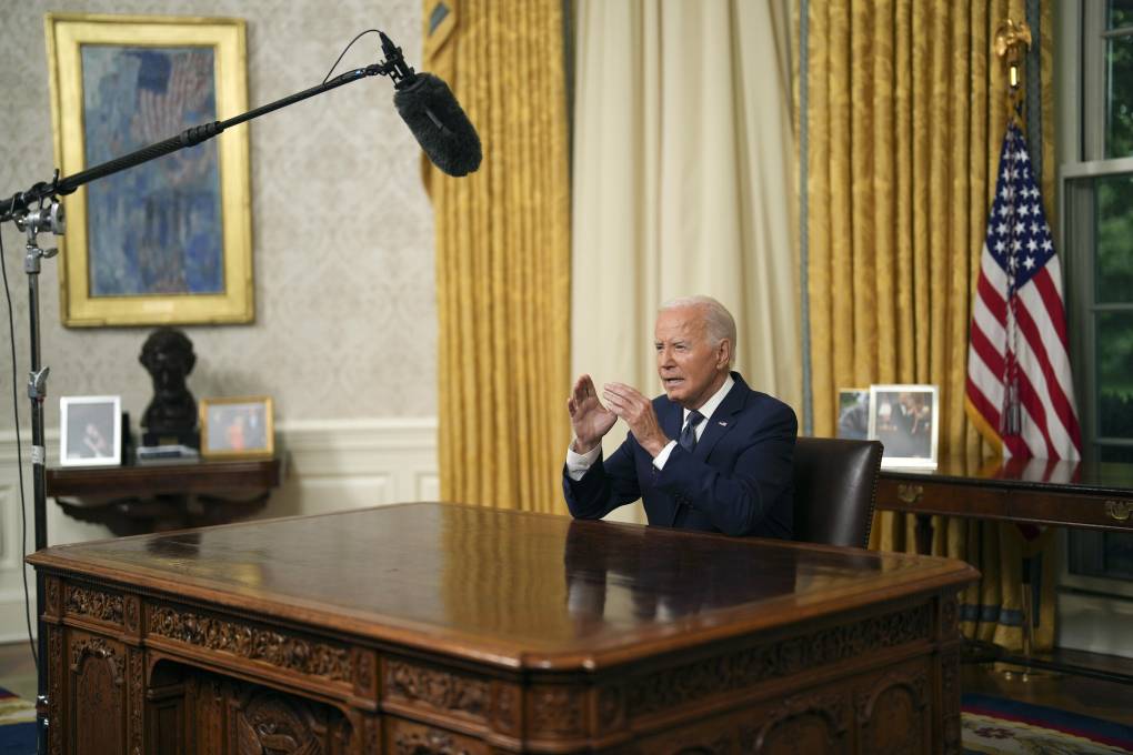 A man in a business suit sits at a desk, gesturing with both hands toward a boom microphone.