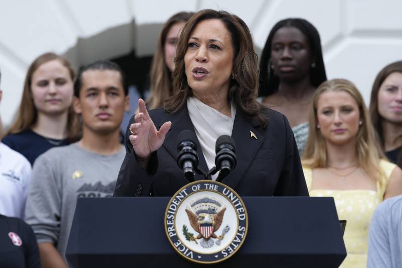 A woman wearing a business suit stands at a podium and gestures with her right hand with several people in the background.