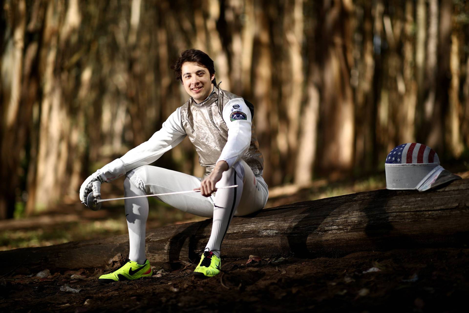 A man wearing fencing gear sits on a log in a wooded area. He is smiling and casually holding a foil.